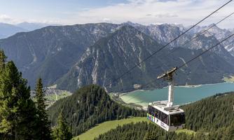 A cable car glides over green hills and a clear lake, surrounded by majestic mountains. The sky is blue and cloudless.