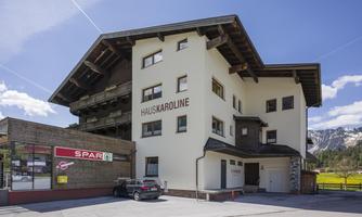 A modern building called "Haus Karoline" with an adjacent supermarket. The surroundings feature a picturesque alpine landscape.