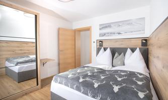 A cozy bedroom with a double bed and modern wooden furniture. The room features a large mirror and beautiful wall decoration.
