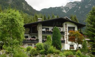 A cozy building named "Rofangarten," surrounded by green trees and beautiful plants. In the background, majestic mountains can be seen.