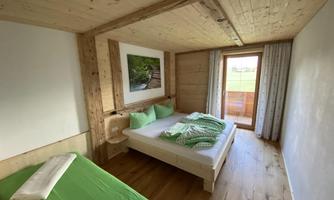A cozy bedroom with wooden walls and a large window. There is a double bed and a single bed, as well as green pillows and blankets.