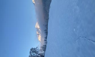 A snowy landscape with gentle hills and a clear blue sky. In the background, snow-capped mountains and a few clouds can be seen.