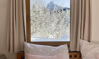 A cozy bedroom with a window through which one can see a snowy forest and mountains. The windows are decorated with bright curtains and the bedding is in soft colors.
