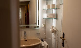 A modern bathroom with a sink and a mirror. The walls are tiled, and there are some shelves with bathroom utensils.