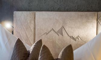 An elegant headboard with a mountain engraving is the focal point of the image. The soft pillows and the modern wall create a cozy atmosphere.