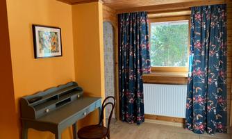 A cozy room with orange walls and floral patterned curtains. A desk and a chair are positioned in front of a window that lets in daylight.