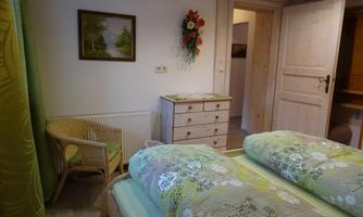 A cozy bedroom with two beds and green bed linens. The room contains a chair, a sink, and a picture on the wall.