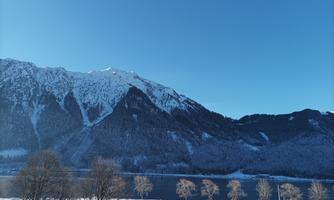 A winter landscape with snow-covered mountains and a clear blue sky. In the foreground, some trees are visible in front of a peacefully flowing body of water.