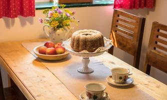 A cozy table with a cake stand, fresh apples, and two cups. In the background, there are windows with red curtains and a bouquet of flowers.