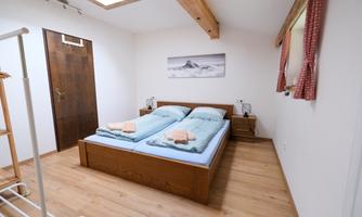 A bright bedroom with a double bed and two nightstands. The walls are painted in soft colors, and there is a window with red curtains.