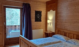 A cozy bedroom with wooden paneling featuring a double bed and access to the balcony. The walls are also made of wood, and there is pleasant lighting.
