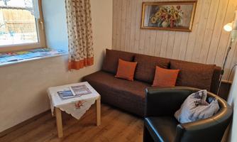 A comfortable living room with a brown sofa and two orange cushions. A side table stands next to it, and a window lets in plenty of light.