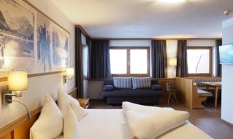 A modern hotel room with a double bed, a sofa, and large windows. The walls are decorated and the atmosphere is cozy.