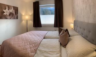 A cozy bedroom with two beds and a warm color scheme. The room has windows with a view outside and soft lighting.