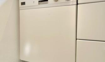 A modern white dishwasher stands in a bright kitchen. Above it is a microwave oven on the countertop.