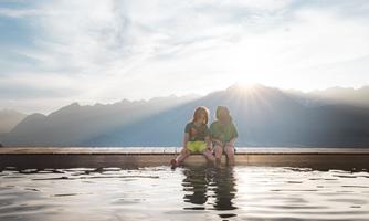 Two children are sitting at the edge of a pool, smiling at the water. In the background, there are majestic mountains and a clear sky with the sun rising.