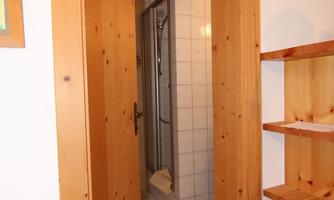 A bathroom with a shower cubicle, visible through a wooden door. The floor is tiled and there is a simple wooden shelf nearby.