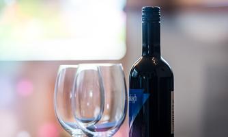 A bottle of red wine stands next to two wine glasses. The background is blurred and features warm colors.