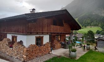 A traditional wooden house with a cozy garden and stacked firewood. In the background, green mountains and a tranquil landscape can be seen.