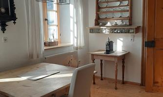 A bright dining area with a wooden table and chairs. On the wall, there is a clock and a shelf with dishes.