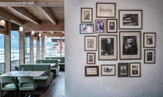 A cozy restaurant with wooden beam ceiling and green seating. Several framed black-and-white portraits hang on the wall.