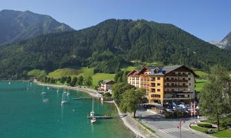 A picturesque lake with boats and a beautiful hotel on the shore. In the background, green mountains and a clear sky can be seen.