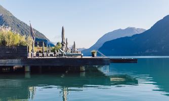 A tranquil lakeside promenade with lounge chairs and sun umbrellas. In the background, majestic mountains and a clear sky can be seen.