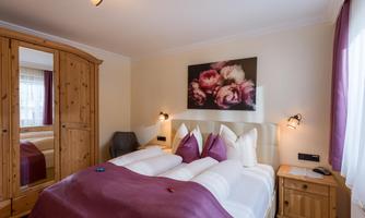A cozy bedroom with a double bed and purple pillows. A large wardrobe and a painted flower painting complete the furnishings.