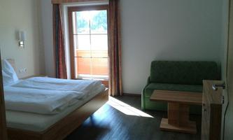 A simple room with a double bed, a sofa, and a table. Natural light streams through the window.
