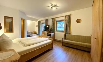 A cozy room with a double bed and a sofa. The decor is bright and friendly, with wooden details and a large window.