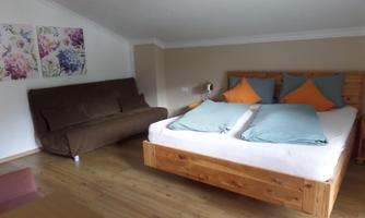 A cozy bedroom with a wooden bed and colorful pillows. In the background, there is a brown sofa bed and decorative pictures on the wall.
