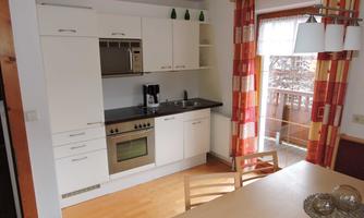 A modern kitchen with white furniture and a dining table.  
Large windows with curtains let in plenty of light.