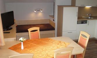 A cozy apartment with a dining table and chairs. In the background, there is a television and a small kitchenette.