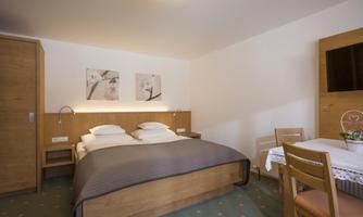 A cozy bedroom with a double bed and wooden furniture. There is a small table with chairs and a TV on the wall.