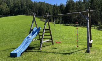 A playground outdoors with a blue slide and a swing. In the background, green meadows and trees can be seen.