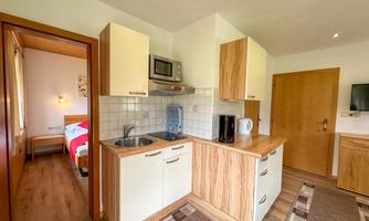 A modern kitchen with wooden cabinets and modern appliances. In the background, there is a bedroom with a red bed.