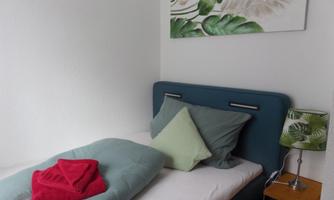 A simple bedroom with a single bed and colorful pillows. On the wall hangs a leaf motif and a bedside table with a green lamp.