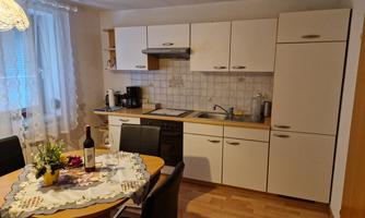 A modern kitchen with white furniture and a round dining table. On the table are flowers and a bottle of wine.