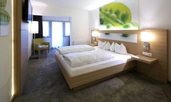 A modern hotel room with a double bed and two bedside tables. The room is bright and friendly, with a cozy armchair and a large window.