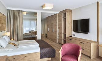 A modern bedroom with wooden furniture and a large window. There is a double bed, a television, and a cozy armchair.