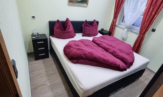 A cozy bedroom with a double bed and purple bedding. The walls are light green and there are red curtains at a window.