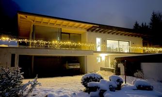 A modern house adorned with Christmas lights at night. The garden is covered in snow, creating a cozy winter atmosphere.