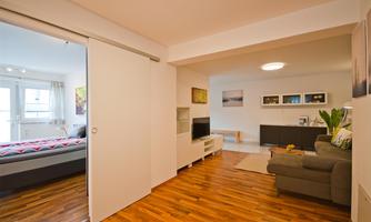 A modern living room with a cozy sofa and a television. On the left side, there is a bedroom with a large bed and plenty of natural light.