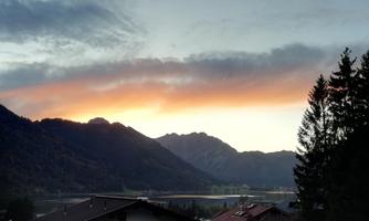 A picturesque mountain landscape at sunset, with warm colors in the sky. In the foreground, roofs of houses and trees are visible.