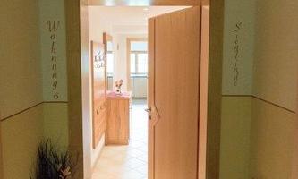 A bright hallway with an open door to a living space. A fire extinguisher is mounted on the wall and a decorative plant is in the foreground.