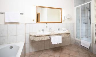 A modern bathroom with two sinks, a large mirror, and a shower. The walls are tiled, and there is a bathtub on the left side.