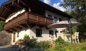 A charming alpine-style house with a wooden balcony. In the foreground, there is a table and chairs under a sun umbrella.