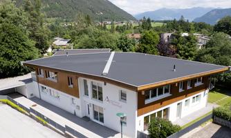 A modern building with a flat roof and wooden elements. Surrounded by green trees and mountains in the background.