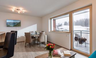 A modern living room with a dining table, chairs, and a TV. Large windows offer a view of the snow-covered landscape outside.