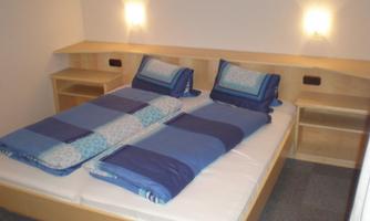 A simple bedroom with a double bed and blue bed linen. There are bedside lamps on the walls and wooden shelves next to them.
